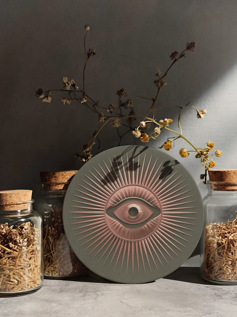Round ceramic coaster featuring a sage green background with a detailed rose gold Evil Eye symbol in the center, surrounded by radiating metallic rays in a sunburst pattern.